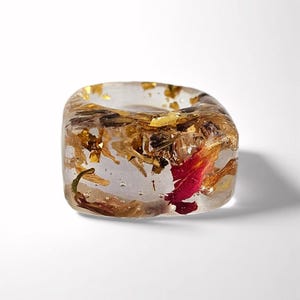 Puede incluir: Anillo cuadrado de resina transparente con bordes redondeados, con pan de oro y flores secas. Un pétalo rojo es visible dentro de la resina. El anillo está sobre un fondo blanco. Diseño único.