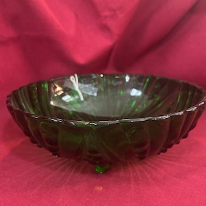 May include: A dark green glass bowl with a textured, ridged design. The bowl has a scalloped edge and sits on three small feet. The background is a solid red color.