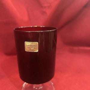 May include: A dark red glass with a clear, stemmed base. The glass has a cylindrical shape and a small, rectangular sticker with gold lettering. The sticker reads "luminarc" and "FRANCE".