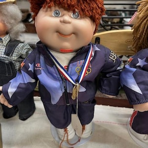 May include: A Cabbage Patch Kid doll wearing a blue and white jacket with stars and the words "USA 1992" on it. The doll is wearing a gold medal around its neck and has red hair.