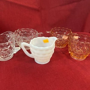 May include: A collection of vintage glass cream and sugar serving pieces. The set includes clear, white, and amber-colored pieces with a diamond pattern. The pieces are arranged on a red surface.