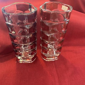 May include: Two clear, faceted glass tumblers with a red hue at the base. The glasses have a geometric design and are displayed on a red surface. The tumblers are empty and appear to be for drinking.