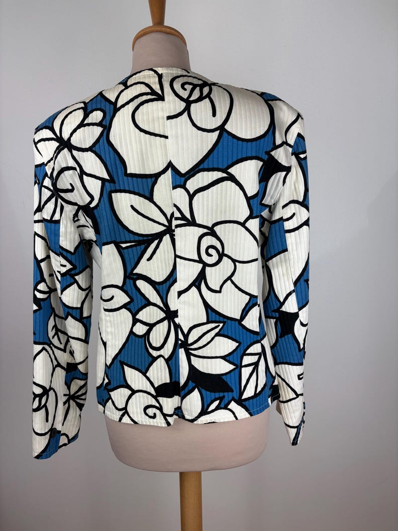 Puede incluir: Una chaqueta de traje de manga larga con estampado floral blanco y azul. La chaqueta tiene una textura acanalada y una silueta ligeramente ajustada.