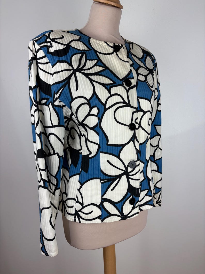 Puede incluir: Una chaqueta con estampado floral azul y blanco con cierre de botones. La chaqueta tiene una textura acanalada y mangas largas.