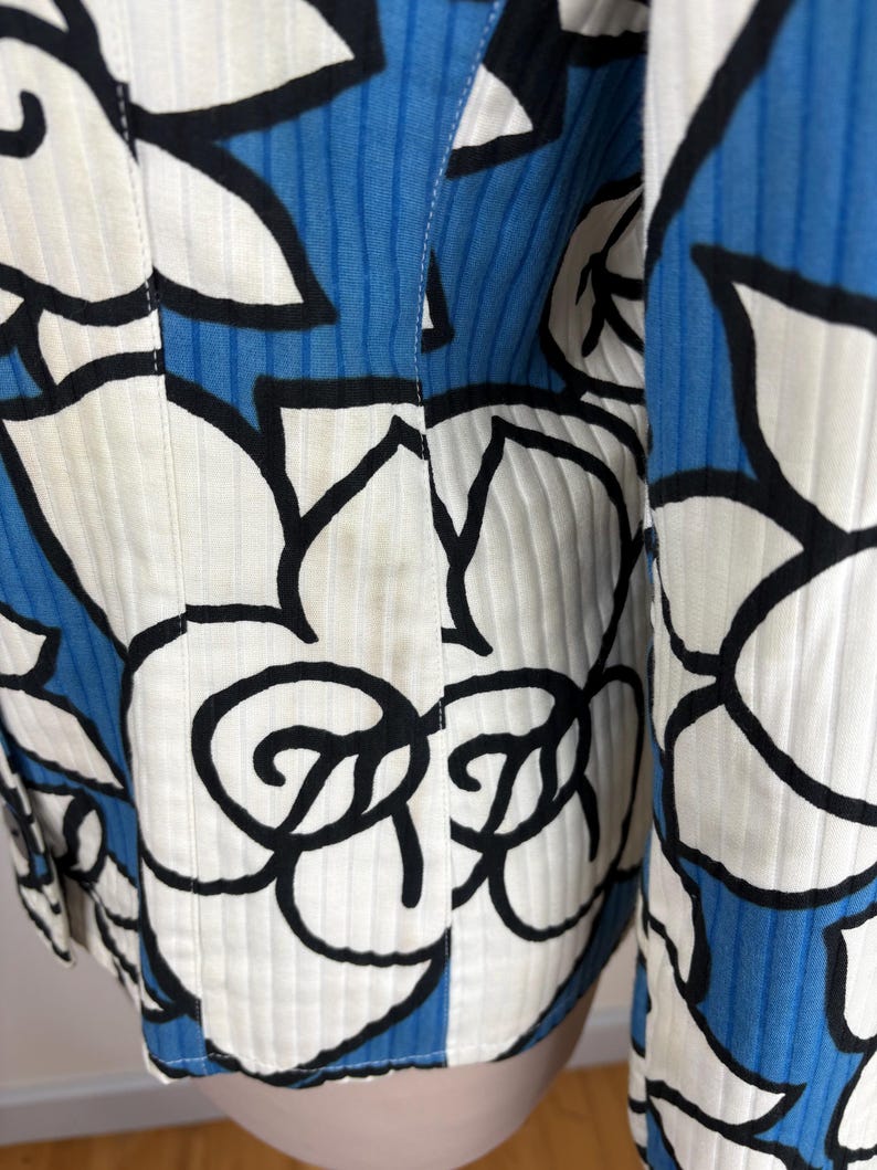 Puede incluir: Una chaqueta con estampado blanco y azul con un dise&ntilde;o floral negro. La chaqueta tiene una textura acanalada y un cierre de bot&oacute;n.