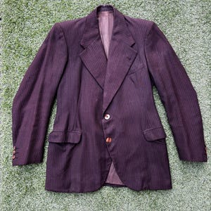 Peut inclure: Un blazer bordeaux foncé à fines rayures verticales. Le blazer a une fermeture à deux boutons, des revers crantés et deux poches avant. Les manches ont trois boutons aux poignets. Le blazer est posé sur une surface verte.