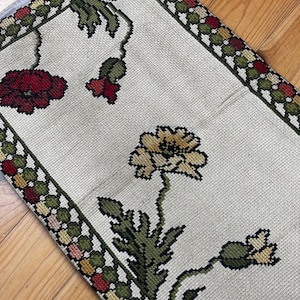 Peut inclure: Tapis rectangulaire crème avec un motif floral. Le tapis présente des fleurs brodées en rouge, jaune et vert, avec une bordure aux couleurs alternées. Le tapis est probablement fait d'une matière tissée.