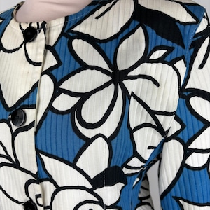 Puede incluir: Una chaqueta con estampado azul y blanco con dise&ntilde;os florales negros. La chaqueta tiene un cierre de botones en la parte delantera y mangas largas.
