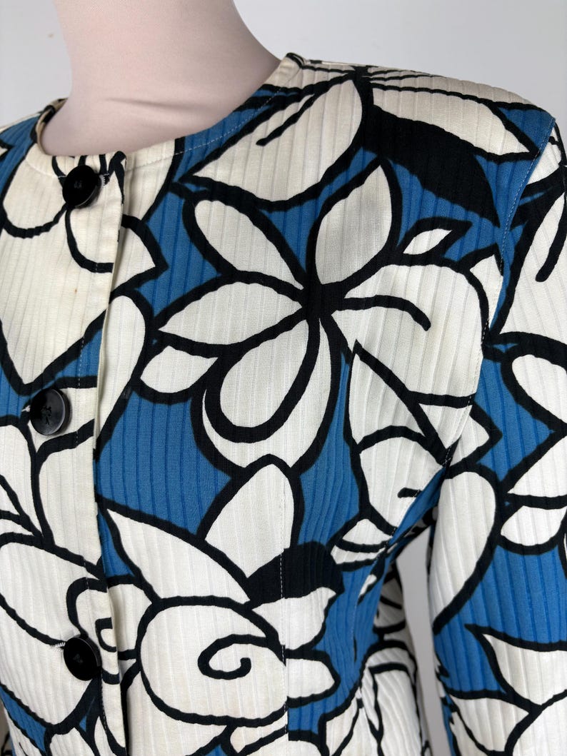 Puede incluir: Una chaqueta con estampado azul y blanco con dise&ntilde;os florales negros. La chaqueta tiene un cierre de botones en la parte delantera y mangas largas.