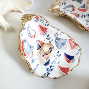 May include: White oyster shell trinket dish with a gold rim and a nautical sailboat pattern in red and blue. A gold ring rests inside the dish. Decorative seashell and another dish are in the background.