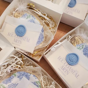 K&ouml;nnte beinhalten: Wei&szlig;e Geschenkboxen mit Seidenpapier, goldenen B&auml;ndern und Artikeln mit floralen Mustern. Jede Box enth&auml;lt einen kleinen Umschlag, der mit einem hellblauen Wachssiegel verschlossen ist. Auf den Umschl&auml;gen steht "LCC Coastal Arts and Studio".