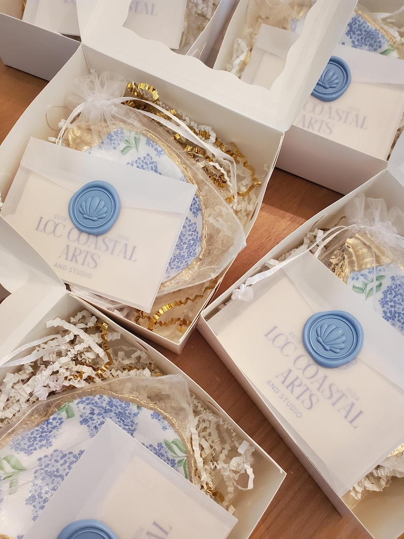 K&ouml;nnte beinhalten: Wei&szlig;e Geschenkboxen mit Seidenpapier, goldenen B&auml;ndern und Artikeln mit floralen Mustern. Jede Box enth&auml;lt einen kleinen Umschlag, der mit einem hellblauen Wachssiegel verschlossen ist. Auf den Umschl&auml;gen steht "LCC Coastal Arts and Studio".