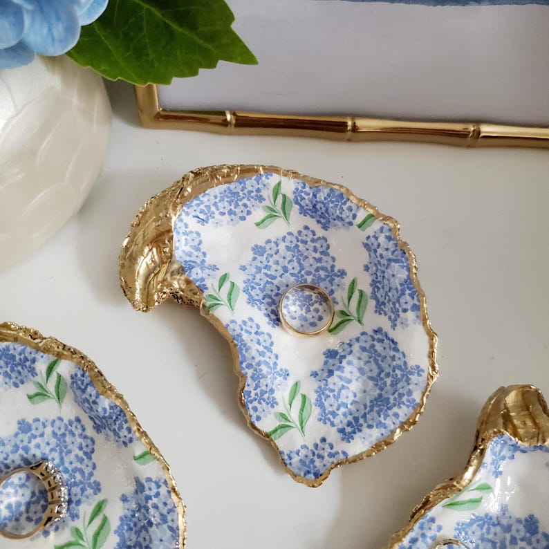 K&ouml;nnte beinhalten: Handbemalte Austernschalen-Ringschale mit goldenem Rand. Die Schale hat ein blaues Hortensien-Blumenmuster und h&auml;lt einen goldenen Ring. Die Schale befindet sich auf einer wei&szlig;en Oberfl&auml;che.