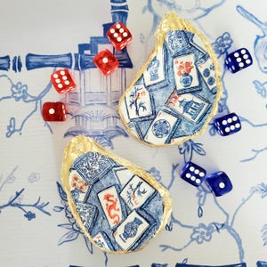 May include: Two decorative oyster shells with gold-painted edges, featuring a decoupage pattern of blue and white mahjong tiles with red accents. The shells are surrounded by several scattered red and blue six-sided dice on a blue and white surface.