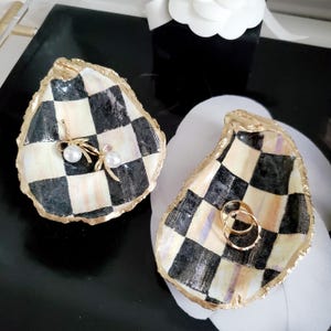 May include: Two decorative oyster shell ring dishes. The shells are painted with a black and white checkered pattern and gold trim. One shell holds a pair of gold earrings with pearls, and the other holds two gold rings.