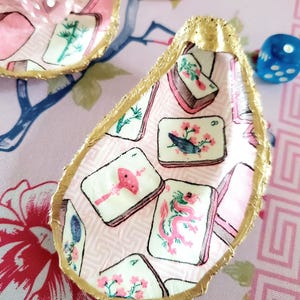 K&ouml;nnte beinhalten: Dekorative Austernschalen mit goldenem Rand, verziert mit einem rosa-wei&szlig;en Mahjong-Fliesenmuster. Die Muscheln sind von rosa W&uuml;rfeln und einem blauen W&uuml;rfel auf einem rosa floralen Hintergrund umgeben.