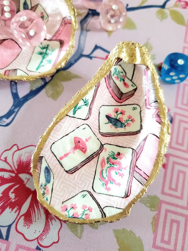 K&ouml;nnte beinhalten: Dekorative Austernschalen mit goldenem Rand, verziert mit einem rosa-wei&szlig;en Mahjong-Fliesenmuster. Die Muscheln sind von rosa W&uuml;rfeln und einem blauen W&uuml;rfel auf einem rosa floralen Hintergrund umgeben.