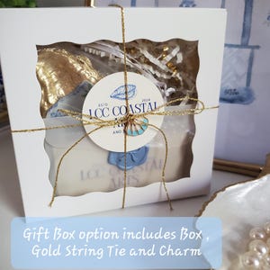 K&ouml;nnte beinhalten: Eine wei&szlig;e Geschenkbox mit einem transparenten Fenster, gebunden mit goldfarbenem Faden. Im Inneren f&uuml;llen goldfarbene Gegenst&auml;nde und wei&szlig;es Papier die Box. Ein rundes Etikett mit dem Text "ICC COASTAL ARTS" und ein blauer Muschelanh&auml;nger sind angebracht.