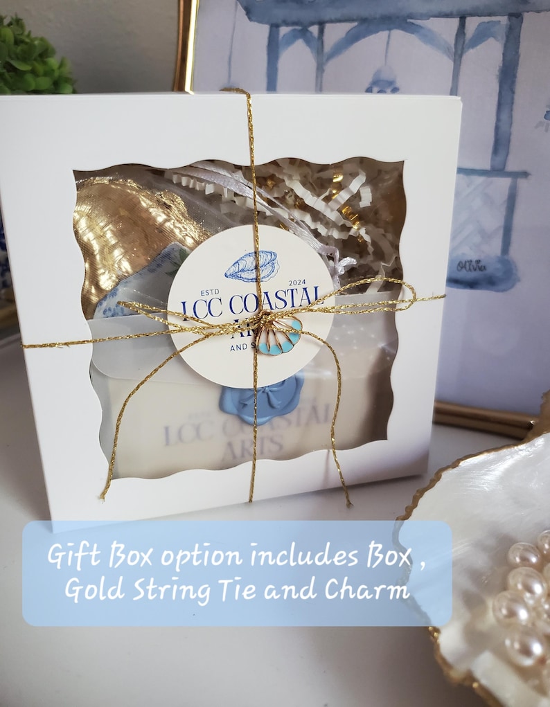 K&ouml;nnte beinhalten: Eine wei&szlig;e Geschenkbox mit einem transparenten Fenster, gebunden mit goldfarbenem Faden. Im Inneren f&uuml;llen goldfarbene Gegenst&auml;nde und wei&szlig;es Papier die Box. Ein rundes Etikett mit dem Text "ICC COASTAL ARTS" und ein blauer Muschelanh&auml;nger sind angebracht.