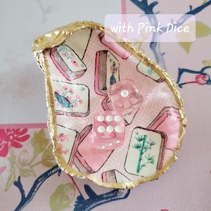 K&ouml;nnte beinhalten: Eine dekorative Austernschale mit goldenem Rand, gef&uuml;llt mit rosa W&uuml;rfeln. Der Innenraum zeigt einen rosa-wei&szlig;en Stoff mit Illustrationen von Blumen und B&uuml;chern. Der Text "with Pink Dice" ist sichtbar.