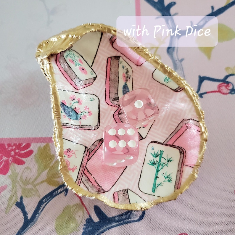 K&ouml;nnte beinhalten: Eine dekorative Austernschale mit goldenem Rand, gef&uuml;llt mit rosa W&uuml;rfeln. Der Innenraum zeigt einen rosa-wei&szlig;en Stoff mit Illustrationen von Blumen und B&uuml;chern. Der Text "with Pink Dice" ist sichtbar.