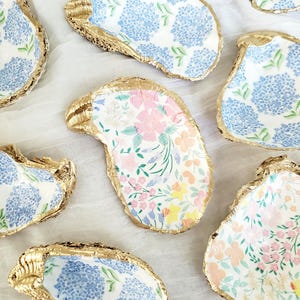 K&ouml;nnte beinhalten: Mehrere dekorative Austernschalen mit goldfarbenen R&auml;ndern. Die Schalen weisen florale Muster in Blau, Rosa und Gelb auf. Sie sind auf einer wei&szlig;en Oberfl&auml;che angeordnet.