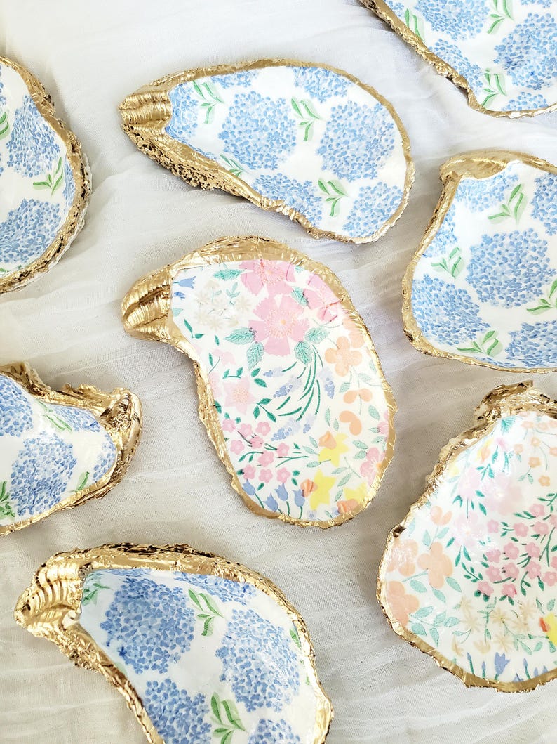 K&ouml;nnte beinhalten: Mehrere dekorative Austernschalen mit goldfarbenen R&auml;ndern. Die Schalen weisen florale Muster in Blau, Rosa und Gelb auf. Sie sind auf einer wei&szlig;en Oberfl&auml;che angeordnet.