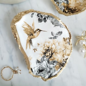 May include: An oval-shaped trinket dish with a gold rim and a white interior. The dish features a hummingbird and floral design in shades of gold, black, and white. A gold ring and pearl earrings are also visible.