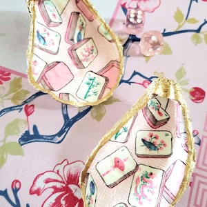 K&ouml;nnte beinhalten: Zwei Austernschalen-Schmuckschalen mit goldfarbenen R&auml;ndern. Das Innere jeder Schale ist mit einem rosa Stoffmuster mit Illustrationen von Mahjong-Steinen verziert. Die Schalen liegen auf einer rosa Oberfl&auml;che mit Blumenmuster.