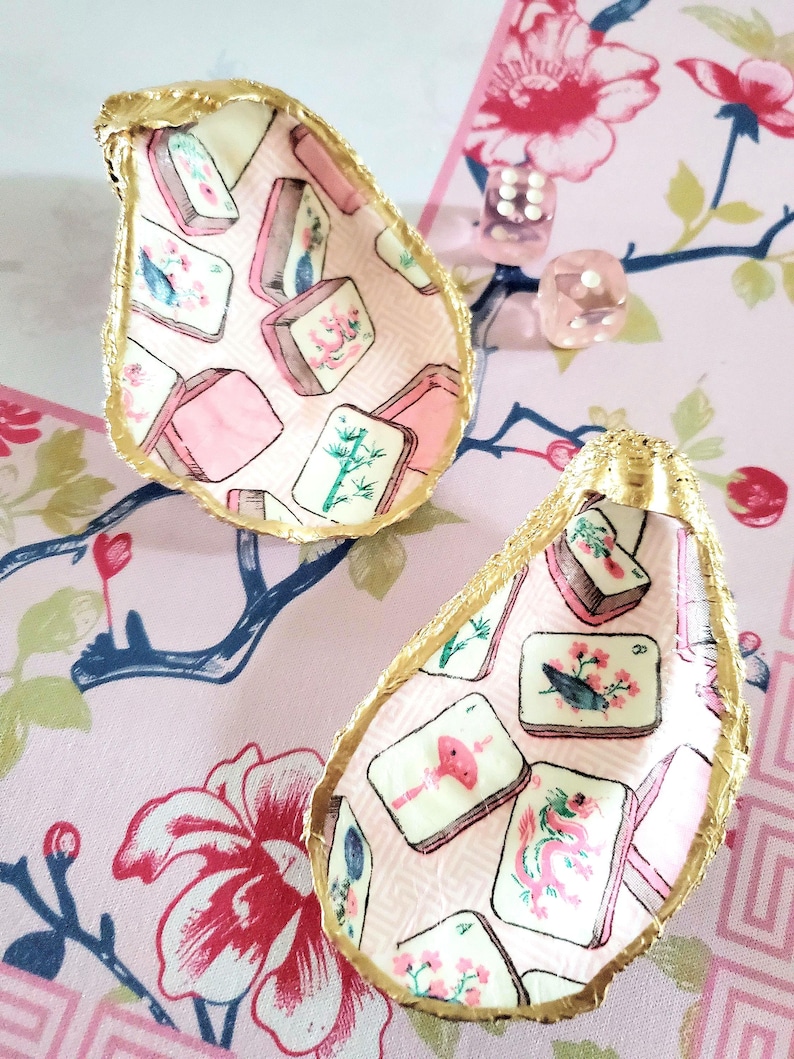 K&ouml;nnte beinhalten: Zwei Austernschalen-Schmuckschalen mit goldfarbenen R&auml;ndern. Das Innere jeder Schale ist mit einem rosa Stoffmuster mit Illustrationen von Mahjong-Steinen verziert. Die Schalen liegen auf einer rosa Oberfl&auml;che mit Blumenmuster.