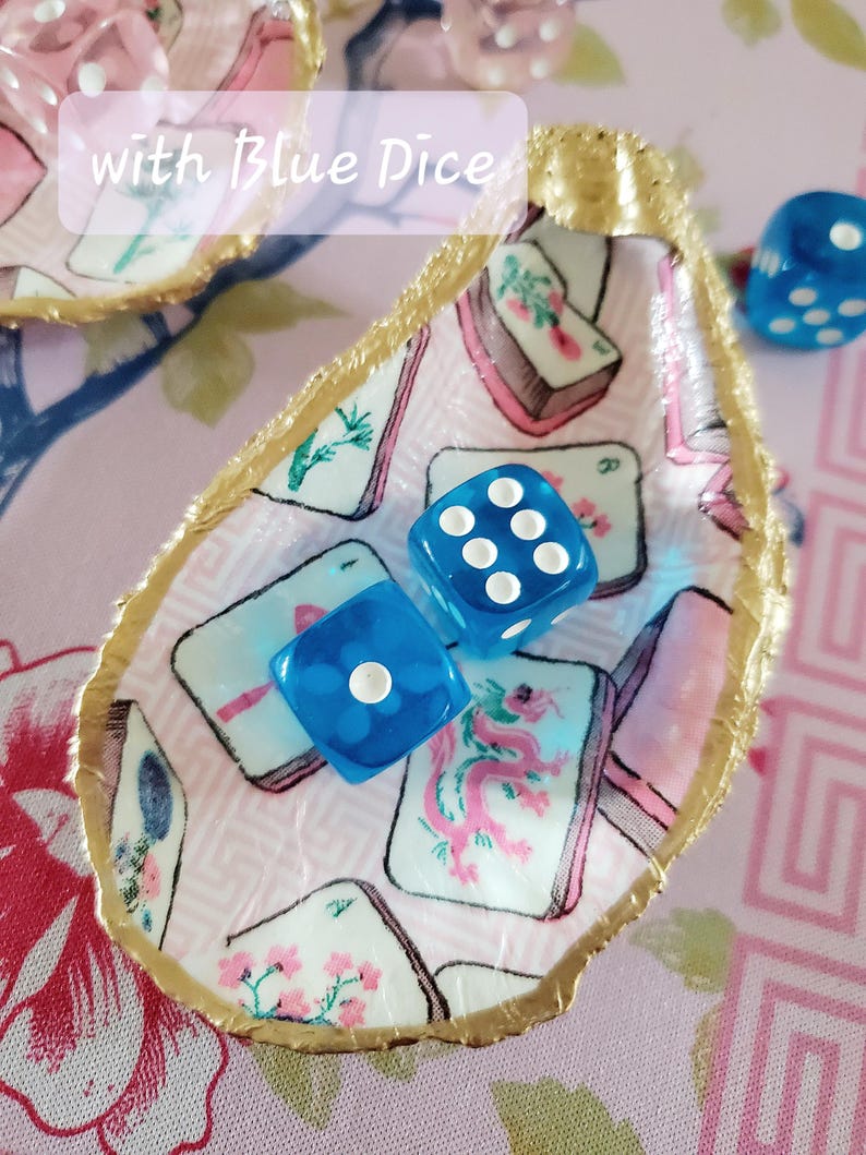 K&ouml;nnte beinhalten: Eine muschelf&ouml;rmige Schmuckschale mit goldfarbenem Rand und einem rosa-wei&szlig;en Muster. Zwei blaue W&uuml;rfel mit wei&szlig;en Punkten liegen darin. Der Text "with Blue Dice" ist sichtbar.