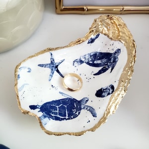 May include: A white ceramic ring dish shaped like an oyster shell with a gold rim. The dish is decorated with blue sea turtle and starfish designs.