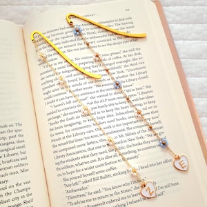 May include: Yellow metal bookmark with a heart-shaped charm featuring the letter "E" and a chain adorned with pearls and blue beads. The bookmark rests on an open book.