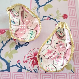 K&ouml;nnte beinhalten: Zwei Austernschalen-Schmuckschalen mit Goldkanten, mit einem rosa-wei&szlig;en Blumenmuster und W&uuml;rfeln. Die Schalen sind mit rosa W&uuml;rfeln gef&uuml;llt und liegen auf einer rosa gebl&uuml;mten Oberfl&auml;che.