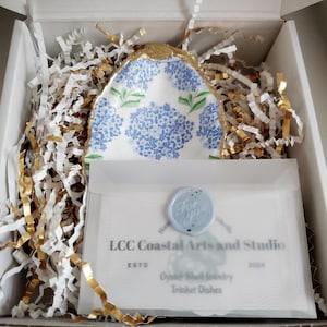 K&ouml;nnte beinhalten: Ein dekoratives Muschelschalen-Schmucksch&auml;lchen mit blauem Blumenmuster und goldenem Rand, verpackt in einer Geschenkbox mit wei&szlig;em und goldenem Crinkle-Papier. Eine Karte mit dem Text "For You" und "LCC Coastal Arts and Studio" ist enthalten.