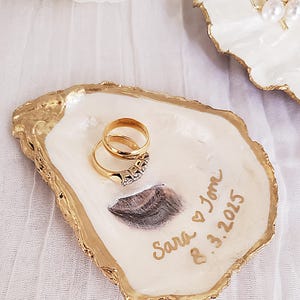 May include: A handcrafted oyster shell ring dish with a gold-painted rim. Two gold wedding rings rest inside. The dish is personalized with the names "Sara & Tom" and the date "8.3.2015" in gold script.