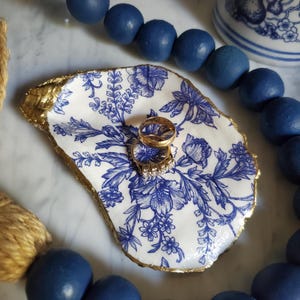 May include: A white ceramic oyster shell dish with a blue and white floral pattern and gold trim. Two gold rings are sitting in the dish.