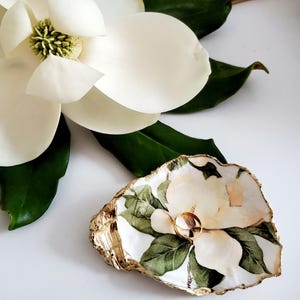 May include: A white oyster shell ring dish with a gold rim and a painted magnolia flower design. A gold ring is sitting in the dish.