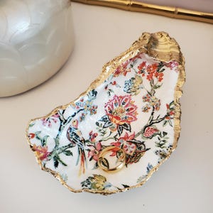 May include: A white ceramic oyster shell dish with a gold rim and a floral pattern. The dish is decorated with a red, yellow, and blue floral design and a small bird. A gold ring is sitting in the dish.