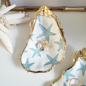 May include: A decorative oyster shell with a gold-painted edge and a white interior featuring blue starfish designs. A gold-colored dragonfly charm with pearl accents is attached to the shell. Other starfish and pearls are in the background.