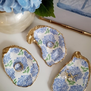K&ouml;nnte beinhalten: Drei Austernschalen-Ringschalen mit goldenem Rand und blauem Hortensienmuster. Eine Schale enth&auml;lt einen Ring, eine weitere einen Ring und die dritte Ohrringe. Im Hintergrund eine Vase mit blauen Hortensien.