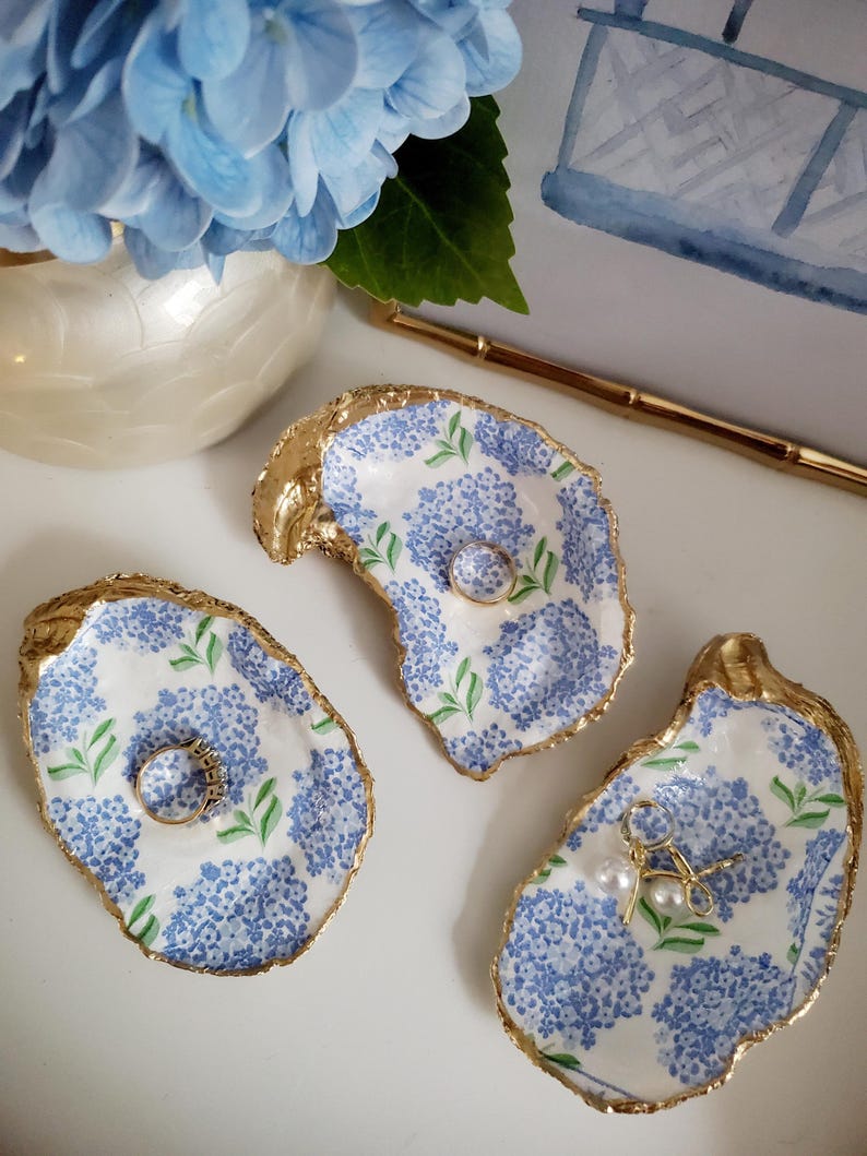 K&ouml;nnte beinhalten: Drei Austernschalen-Ringschalen mit goldenem Rand und blauem Hortensienmuster. Eine Schale enth&auml;lt einen Ring, eine weitere einen Ring und die dritte Ohrringe. Im Hintergrund eine Vase mit blauen Hortensien.