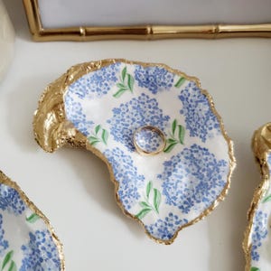 K&ouml;nnte beinhalten: Eine Ringablage aus Austernschale mit goldenem Rand und blauem Blumenmuster. Ein goldener Ring liegt in der Schale. Die Schale ist wei&szlig; mit blauen Hortensienbl&uuml;ten und gr&uuml;nen Bl&auml;ttern.