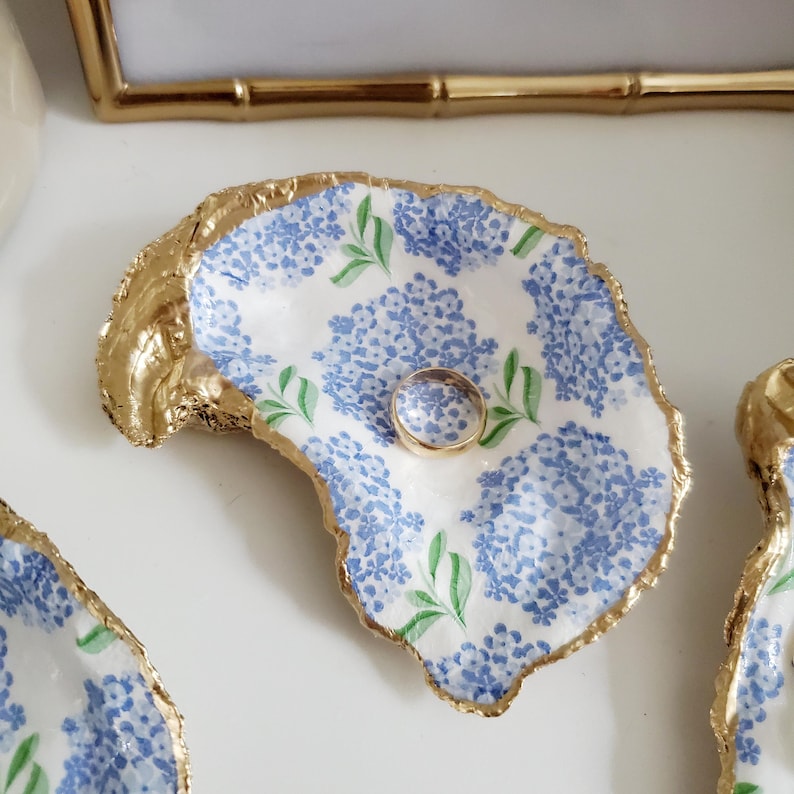 K&ouml;nnte beinhalten: Eine Ringablage aus Austernschale mit goldenem Rand und blauem Blumenmuster. Ein goldener Ring liegt in der Schale. Die Schale ist wei&szlig; mit blauen Hortensienbl&uuml;ten und gr&uuml;nen Bl&auml;ttern.