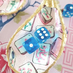 K&ouml;nnte beinhalten: Dekorative Austernschale mit goldfarbenem Rand, gef&uuml;llt mit blauen W&uuml;rfeln. Das Innere der Schale zeigt ein rosa-wei&szlig;es Muster mit Spielkartenillustrationen. Weitere W&uuml;rfel sind im Hintergrund zu sehen.