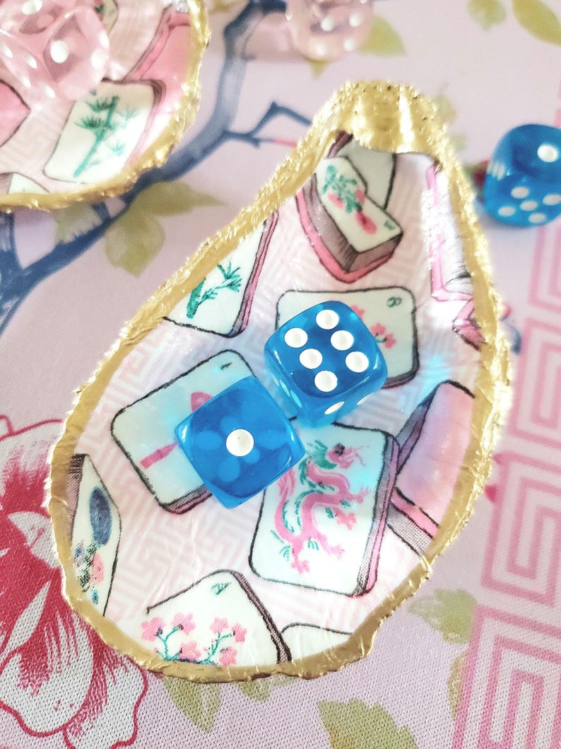 K&ouml;nnte beinhalten: Dekorative Austernschale mit goldfarbenem Rand, gef&uuml;llt mit blauen W&uuml;rfeln. Das Innere der Schale zeigt ein rosa-wei&szlig;es Muster mit Spielkartenillustrationen. Weitere W&uuml;rfel sind im Hintergrund zu sehen.
