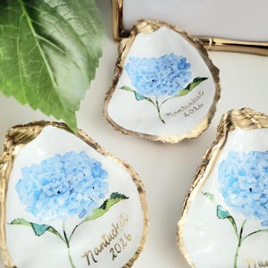 May include: Three white oyster shells with gold rims, each hand-painted with a blue hydrangea flower. The shells have the word "Nantucket" and the year "2026" written in gold script. The shells are arranged on a white surface.