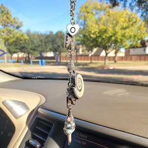 May include: A car charm with a seashell theme. The charm features a heart-shaped pendant with the letter "O", seashells, and a large circular pendant with a mother-of-pearl design. The charm is suspended from a chain.