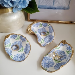K&ouml;nnte beinhalten: Drei dekorative Ringhalter aus Austernschalen. Jede Schale ist mit einem blauen Hortensienbl&uuml;tenmuster bemalt und mit Gold verziert. Ringe und Perlen werden in den Schalen ausgestellt. Im Hintergrund steht eine Vase mit blauen Hortensien.