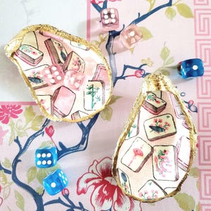 May include: Two decorative oyster shell dishes with gold trim, filled with pink and white Mahjong tiles. Scattered around are clear and blue dice, all set against a floral patterned background.